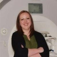 Headshot of Christina Webb with long brown hair, green blouse, and black sweater, standing in front of an MRI machine.