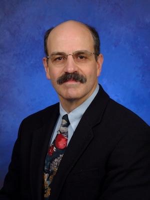 Headshot of Vernon Chinchilli with mustache, glasses, light blue shirt, multi-colored tie, and black jacket.