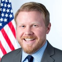 Michael Donovan head shot in gray suit, blue shirt and tie, and flag background.