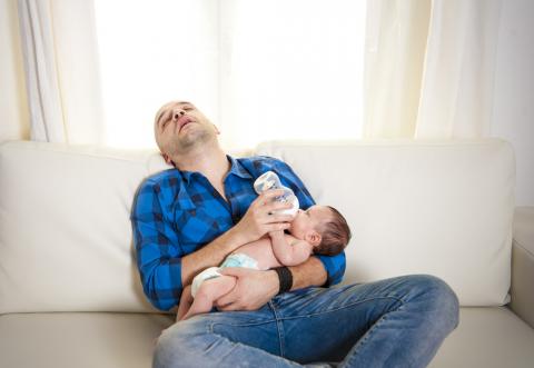 Photo of a new dad sleeping on a couch while feeding a baby.