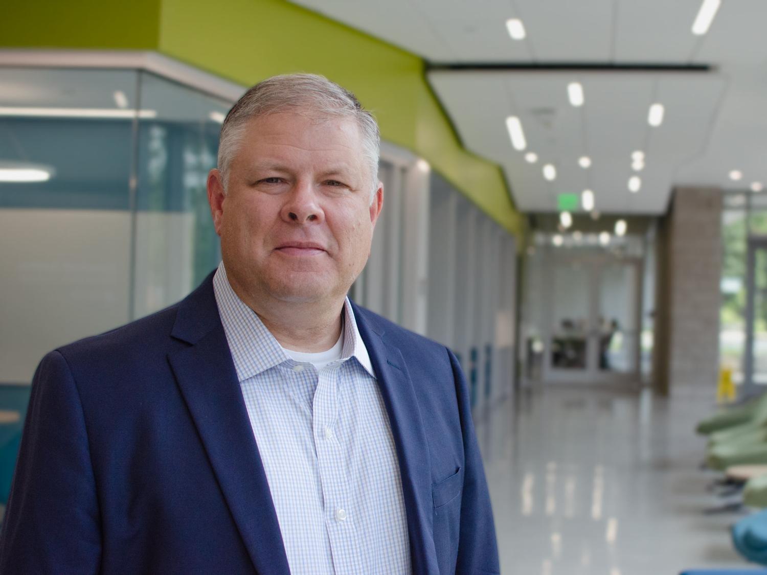 Dennis Scanlong headshot with navy blazer, light blue button-down shirt , grey hair in building with white, gray, and bright green walls and white floor.