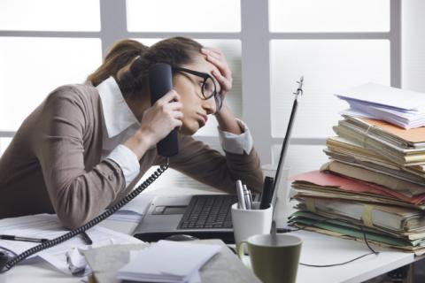 Photo of a woman sitting in front of a laptop, with stack of papers, talking on the phone, and leaning her head on her hand.