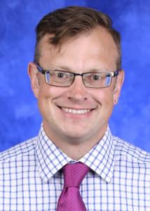 Headshot of Wesley Raup-Savage, a white man with short, brown hair and glasses wearing a blue checkered shirt and pink tie.