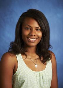Headshot of Ashley Patterson in front of a blue background with dark, curly hair wearing a necklace and tank top.