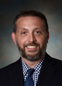 Headshot of Christian Connell with a beard, white and navy checkered shirt, navy tie, and black jacket.