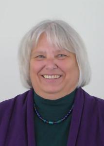 Headshot of Cynthia Mitchell with mid-length grey hair, green turtleneck, purple cardigan and matching necklace.