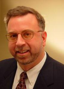 Headshot of David Abler wearing round glasses suit and tie in front of a light-colored wall. 