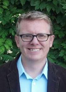 Headshot of Derek Kreager outside with glasses and light-colored hair wearing a blue dress shirt and black suit jacket 