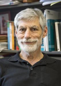 Headshot of Eric Plutzer in front of a book shelf with grey hair and beard wearing a black, collared shirt. 