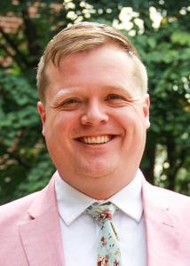 Headshot of Joel Landry outside with short, blonde hair wearing a pink suit jacket and floral tie.