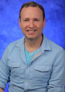 Headshot of Joshua Muscat in front of a blue background with short dark hair wearing a light blue button-up. 