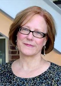 Headshot of Linda Wray with mid-length red hair wearing glasses statement earrings and patterned shirt.