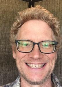 M. Luke Smith head shot with black framed glasses and blond curly hair smiling.