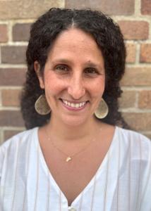 Caucasian woman with long curly hair pulled back and parted in the center, hazel eyes, wearing drop gold earrings and a friendly smile.