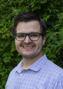 caucasian male with short brown hair, hazel eyes, friendly smile, clean shaven, wearing glasses and plaid button down shirt soft blue colours