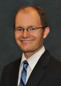 Headshot of Daniel Mallison with glasses, blue striped tie, and black suit jacket.