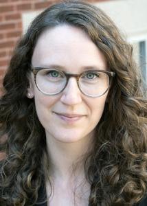 Headshot of Nicole Kreisberg, a white woman with long, brown, curly hair and glasses wearing a black jacket.