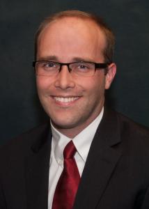 Headshot of Chris Whipple, a white man with glasses, very short blonde hair, and a black suit with red tie.