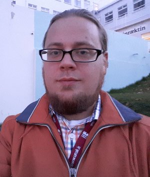 Headshot of Evan with brown hair, goatee, glasses, plaid shirt, and orange jacket.