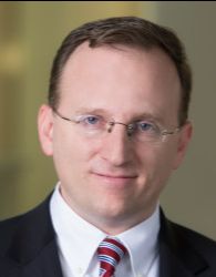 Headshot of Conrad Hackett with brown hair, glasses, white shirt, red and white striped tie, and black jacket.