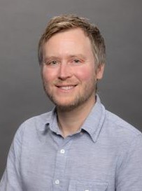 Caucasian man with green eyes, short blonde hair, friendly smile, wearing light blue button-up shirt