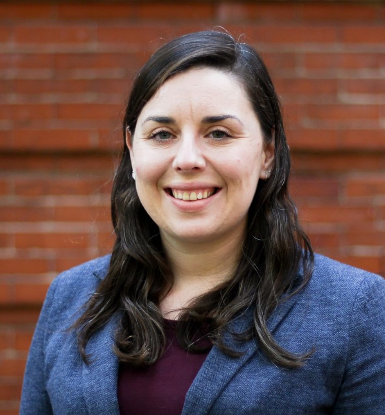 Caucasian woman with long brown hair, blue eyes and friendly smile; wearing blue herringbone suit and burgundy blouse