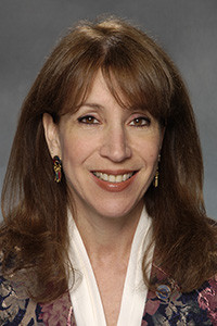 Headshot of Diana with long brown hair, white blouse, and multi-colored jacket.