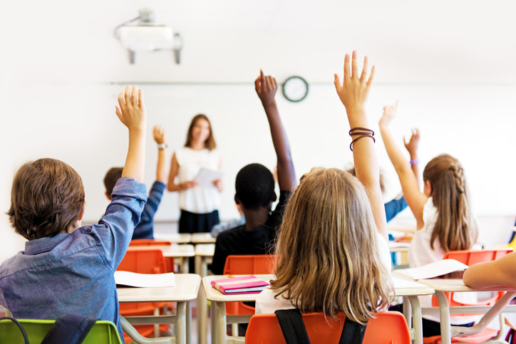 Photo of students in a classroom, raising their hands.