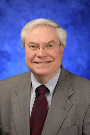 Headshot of Raymond with gray hair, glasses, light blue shirt, red tie, and gray jacket.