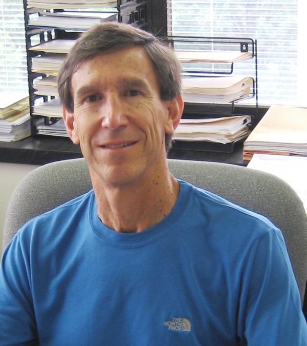 Photo of Dr. Barrett Lee with short brown hair and blue shirt, sitting in a gray office chair in front of a window.