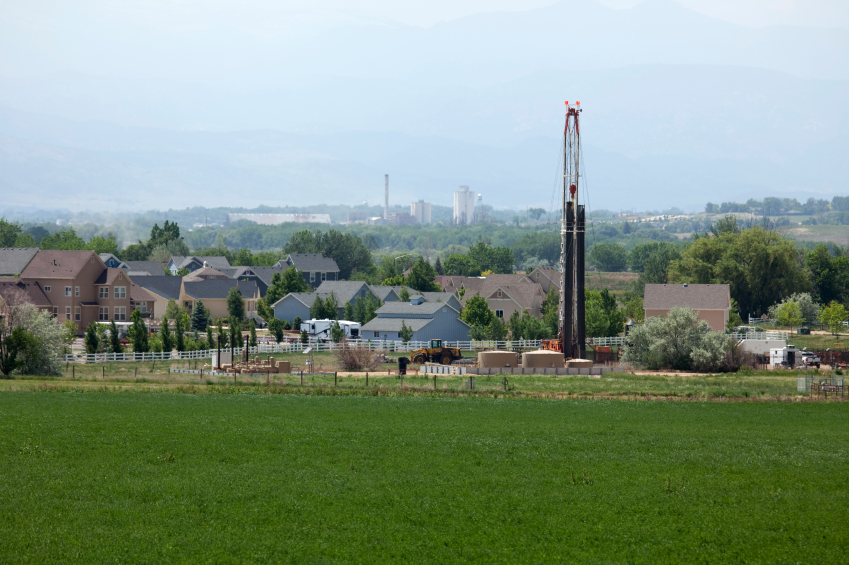 Photo of a gas well with a green open field in front of it and a community behind it.