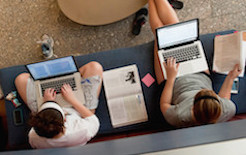 Photo of two students sitting on a bench with their laptops and textbooks while studying.