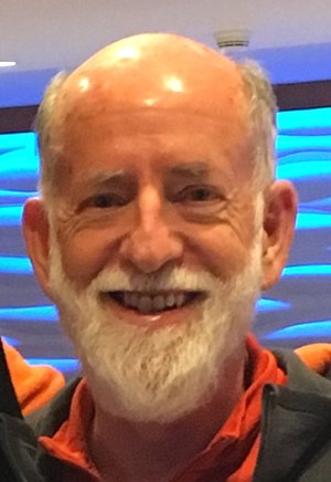 Headshot of Andrew with white hair, beard, orange shirt, and gray jacket.