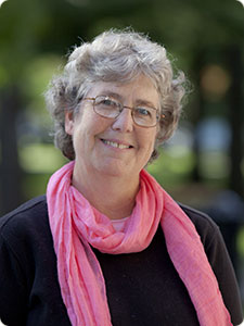 Headshot of Nancy Riley with gray hair, glasses, pink scarf, and black shirt.