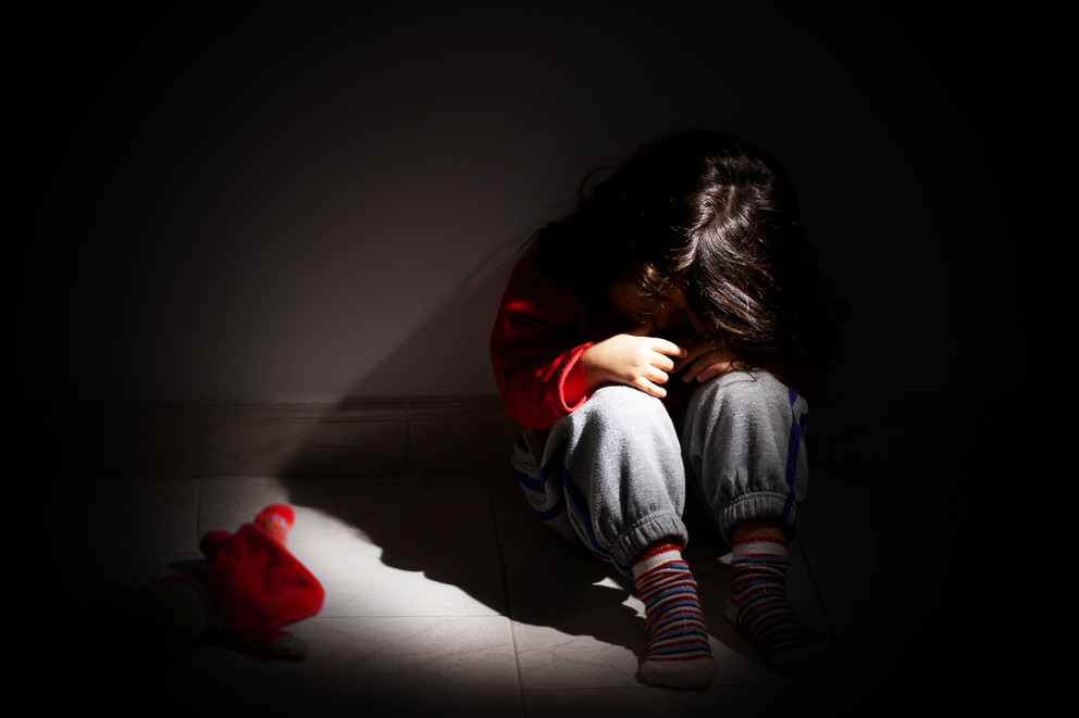 A child sitting against a wall in the fetal position in a darkened room.