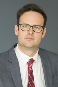 Headshot of Max Crowley with short dark hair, glasses, white shirt, red and gray striped tie, and gray jacket.