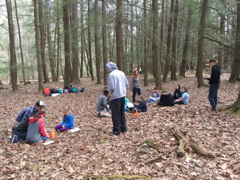 An instructor talking with groups of children who are sitting in a clearing in the woods.