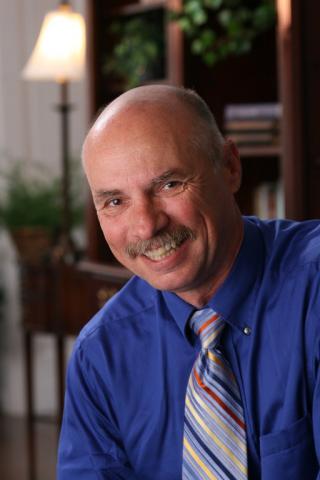 Headshot of Neil Sharkey with mustache, blue shirt, and multi-color striped tie.