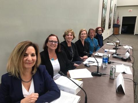 Six women sitting at a long table with microphones and stacks of paper in front of them.