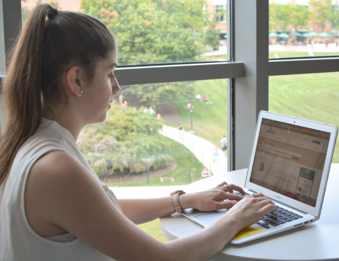 A female student sitting in front of a laptop, with the University Park campus seen below through a window.
