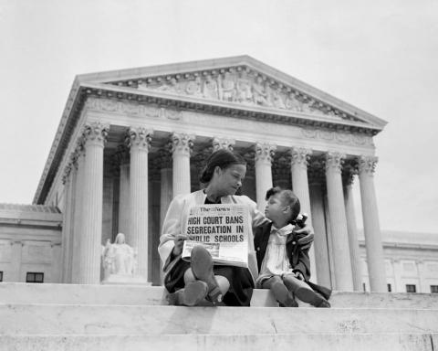 An African American mother and daughter sit on the steps of the US Supreme Court Building holding a newspaper announcing banned segregation.