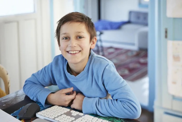 Young boy smiling in front of a computer with long sleeve, blue shirt and short brown hair.