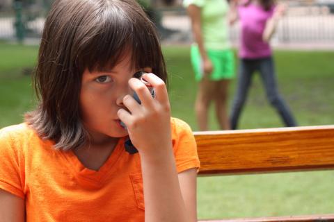 A little girl sitting outside on a bench and using an inhaler.