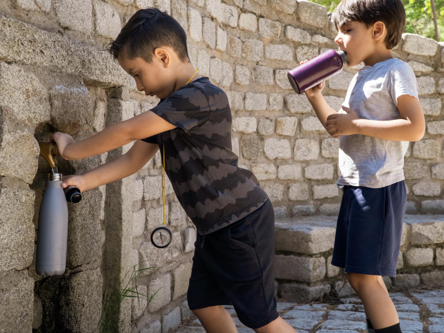 Two young Hispanic children filling water bottles at an outside water fountain.