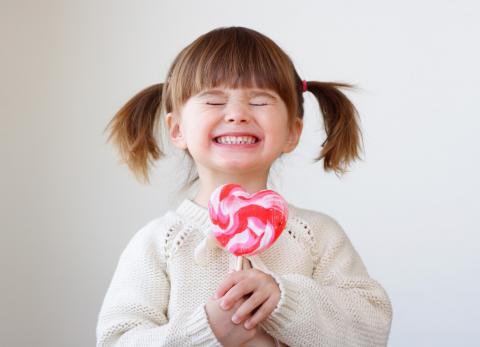 Little girl with her hair in pigtails, holding a large heart sized lollipop, and with a big smile on her face.