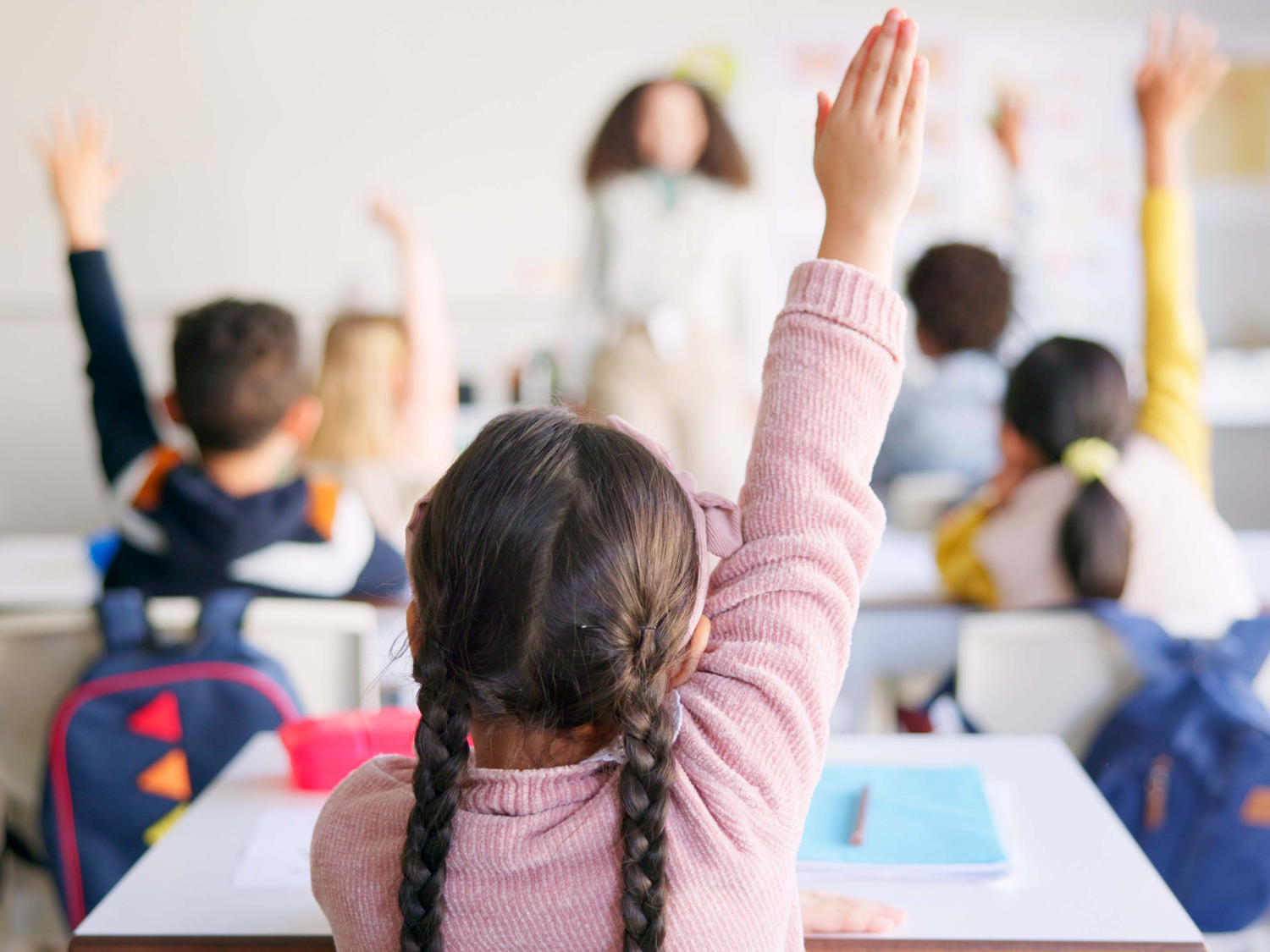 Young girl with braids and pink shirt raises her hand in a classroom.