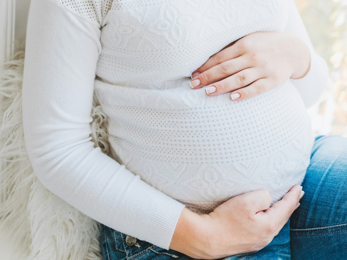 Pregnant woman in white, long sleeved shirt and jeans sitting down holding her belly.