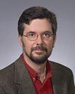 Headshot of Kai Schafft with dark brown hair, glasses, goatee, red shirt, and brown jacket.