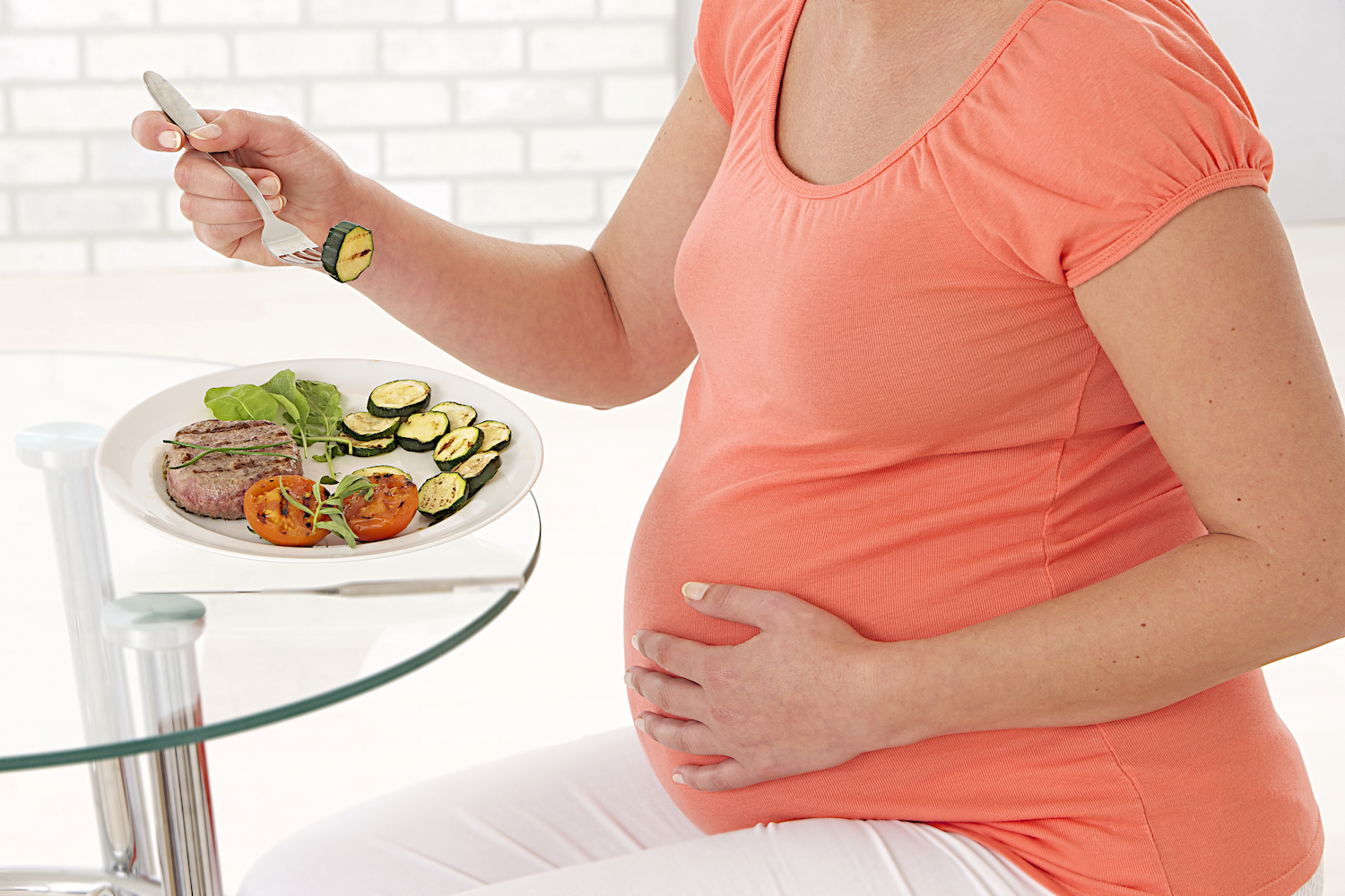 Pregnant woman wearing bright orange shirt sitting at a table and eating salad and a steak.