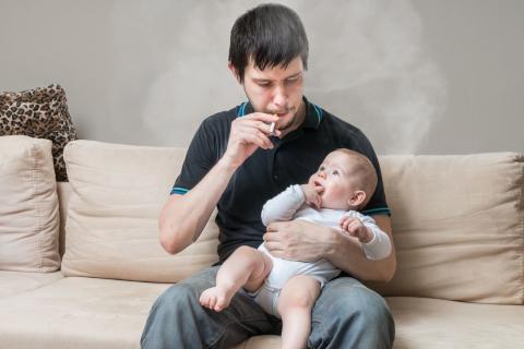A man sitting on a couch, smoking a cigarette, and holding a baby.